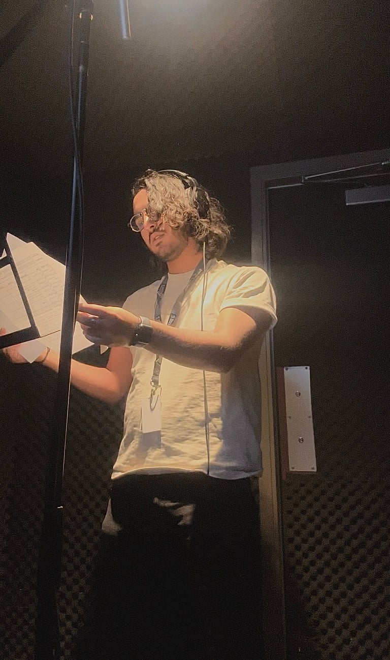 Person with curly blonde hair and glasses standing at a microphone in a recording studio, wearing a yellow shirt and holding a script