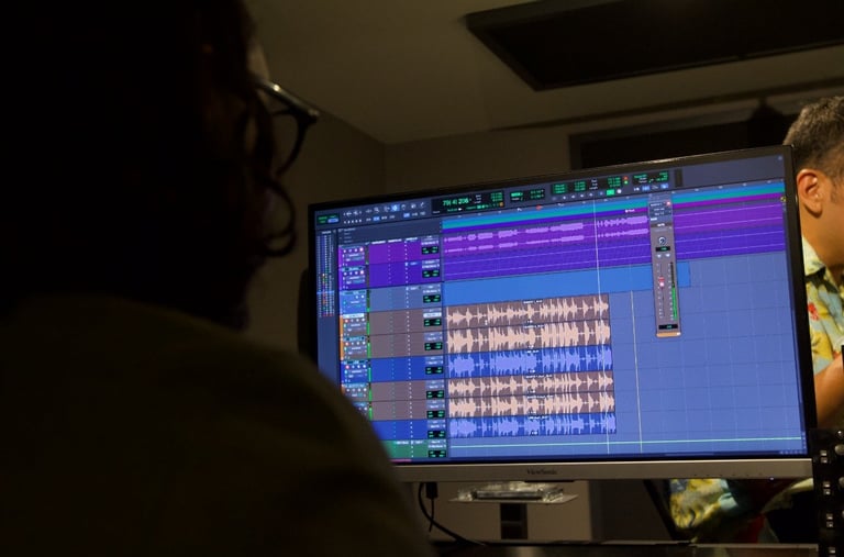 Two people viewing a computer monitor displaying digital audio waveforms in a music production software interface with colorful tracks and controls